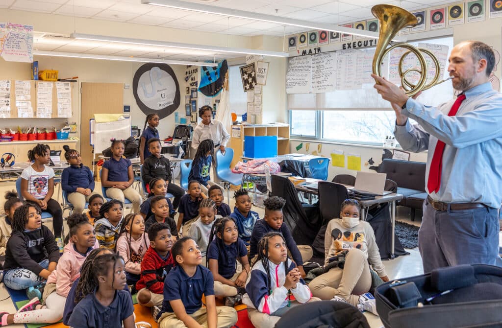 A classroom of school kids listen as a man blows a horn