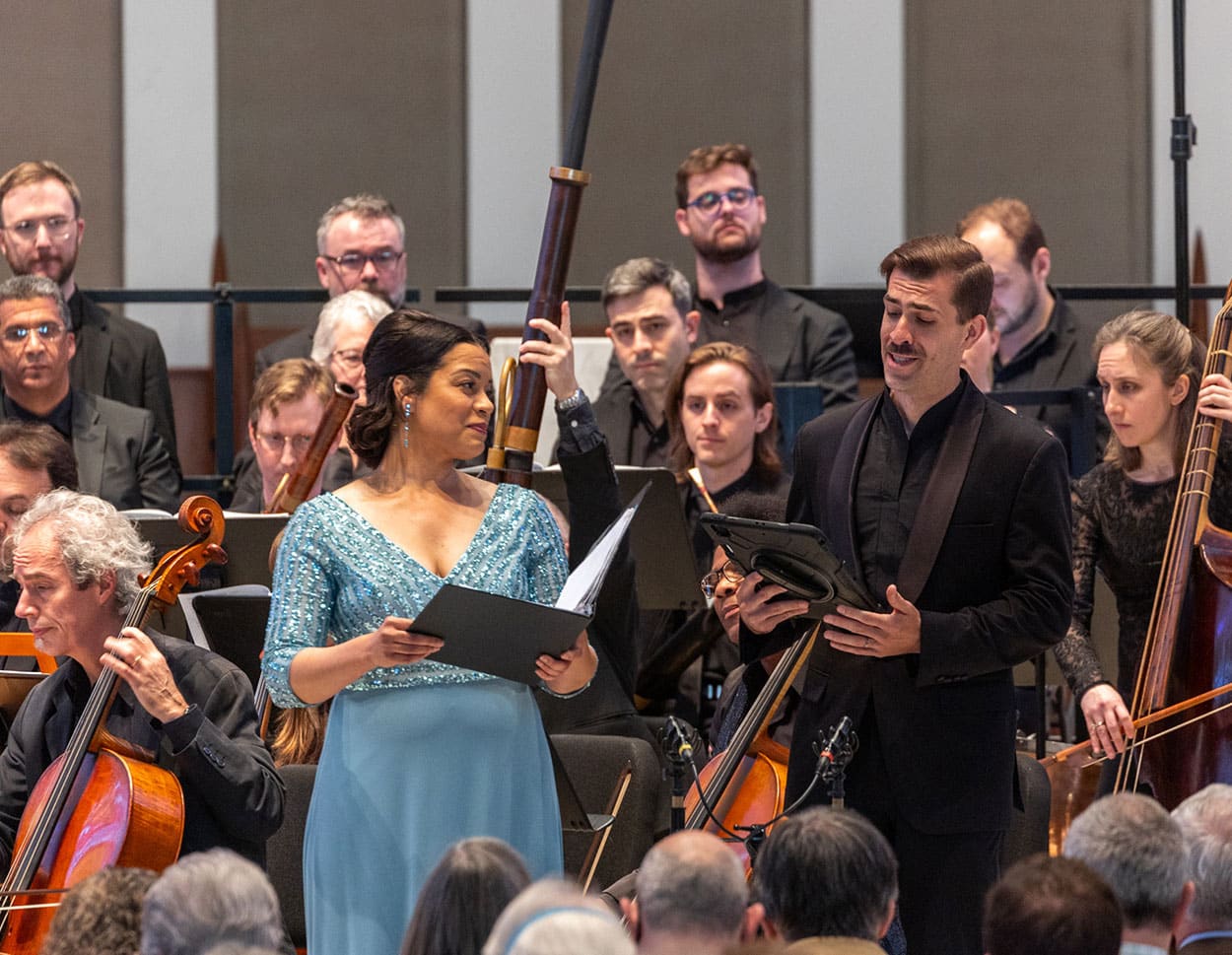 A woman in a blue dress and a man in a tuxedo singing a duet with the orchestra in the background